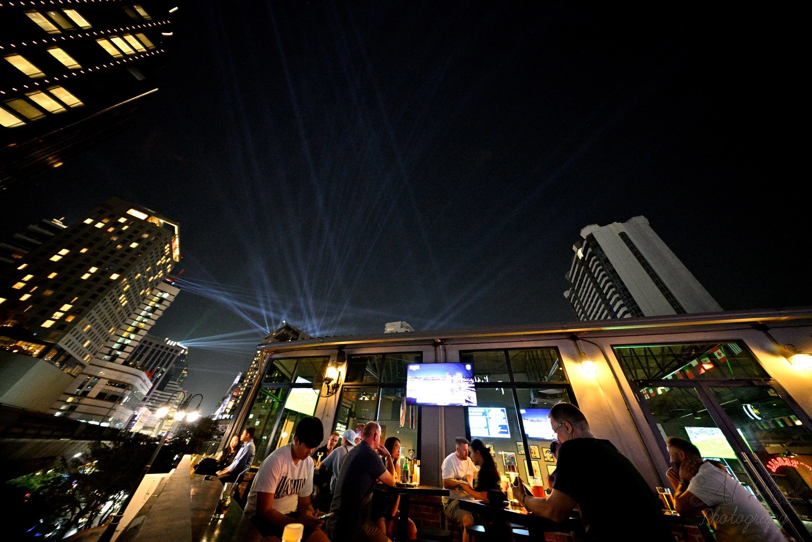 Nighttime view of guests enjoying drinks at the outdoor rooftop deck of Upper Deck Sports Bar, set against the illuminated Bangkok city skyline with light beams shining into the night sky.