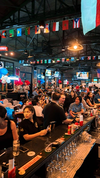 A lively and crowded sports bar interior with patrons sitting at the bar counter and tables. The ceiling is decorated with international flags, and multiple TV screens are broadcasting sports in the background.