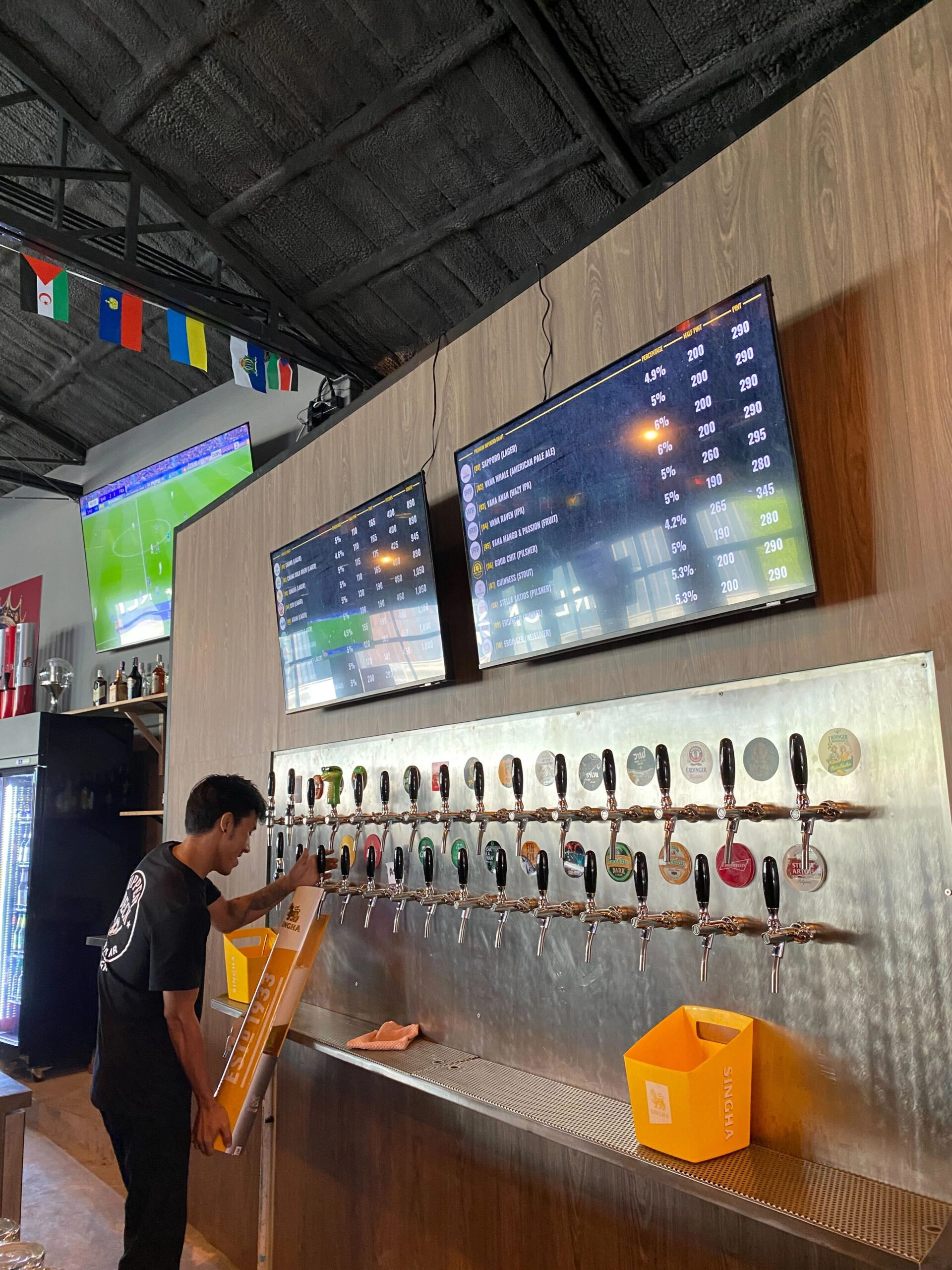 A staff member setting up at the long stainless steel draft tap wall, featuring over 20 craft beer taps mounted below digital screens displaying the current draft beer menu and prices.